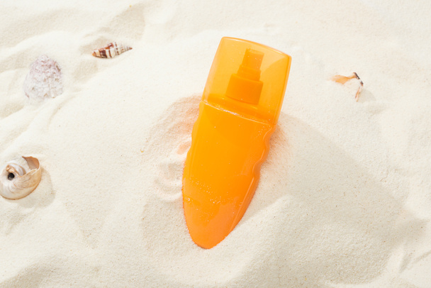 оранжевая бутылка солнцезащитного крема в песке с ракушками
 - Фото, изображение
