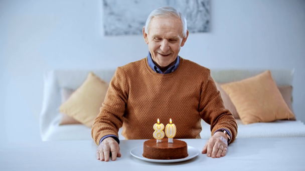 happy senior man looking at birthday cake with candles and smiling at home  - Кадры, видео