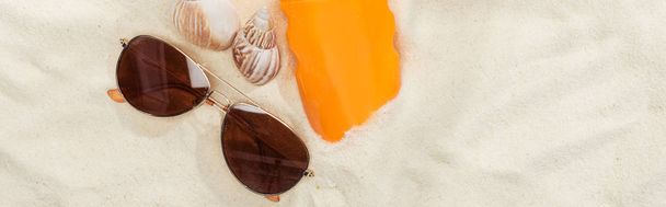 pomarańczowa butelka z filtrem przeciwsłonecznym na piasku w pobliżu muszli i okulary przeciwsłoneczne, panoramiczny strzał - Zdjęcie, obraz