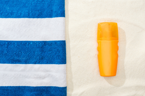 top view of sunscreen and striped blue and white towel on golden sand - Foto, afbeelding
