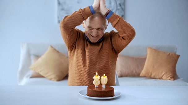 šťastný starší muž, který se dívá na narozeninový dort s hořící svíčkami, směje se a gestikuloval doma  - Záběry, video