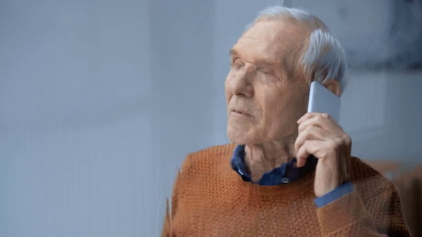 selective focus of senior man talking on smartphone near window with pouring rain  - Metraje, vídeo
