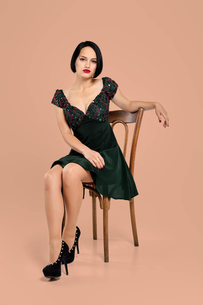 Gorgeous lady in short dress sitting on chair in studio with beige background - Photo, Image