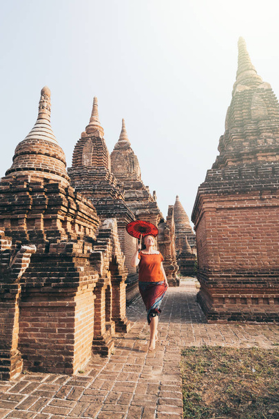 Blonde Caucasian woman with red umbrella runs among temples and pagodas of ancient Bagan in Myanmar - Photo, Image