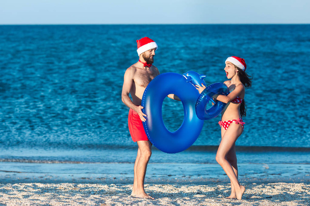 bärtige Kerle und Mädchen mit Weihnachtsmannhüten spielen mit aufblasbaren Kreisen und einem aufblasbaren Delphin am Meeresstrand. - Foto, Bild
