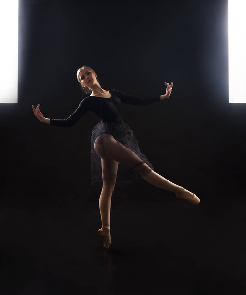 Chica encantadora bailarina en un traje negro, está bailando un ballet a la luz del contorno. Disparando una actuación sobre un fondo oscuro. - Foto, Imagen