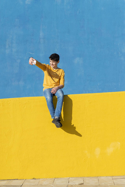 Front kilátás egy fiatal fiú visel alkalmi ruhák ül egy sárga kerítés ellen, a kék fal, miközben egy selfie a smartphone - Fotó, kép