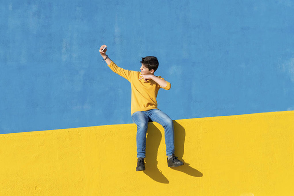 Vista frontal de un niño joven con ropa casual sentado en una valla amarilla contra una pared azul mientras toma una selfie con un teléfono inteligente
 - Foto, Imagen