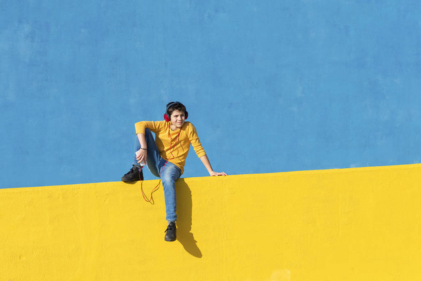 Vooraanzicht van een jonge jongen dragen casual kleding zittend op een gele hek tegen een blauwe muur terwijl het gebruik van een mobiele telefoon naar luisteren muziek door koptelefoon - Foto, afbeelding