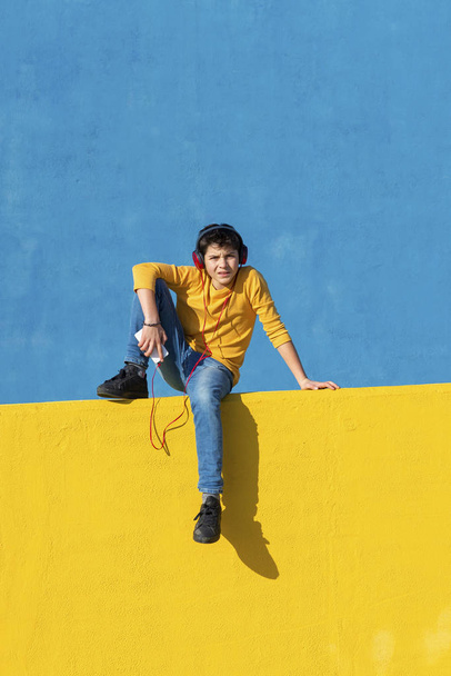 Вид спереди на мальчика в повседневной одежде, сидящего на жёлтом заборе у синей стены, используя мобильный телефон для прослушивания музыки с помощью наушников
 - Фото, изображение