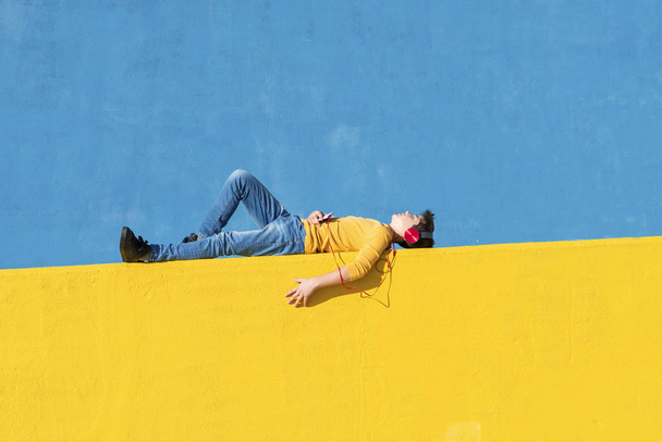Front view of a young boy wearing casual clothes lying on a yellow fence against a blue wall while using a mobile phone to listening music by headphones - Foto, afbeelding