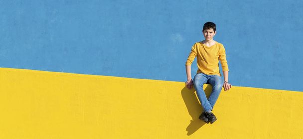 Vista frontal de un niño joven con ropa casual sentado en una valla amarilla contra una pared azul en un día soleado
 - Foto, imagen