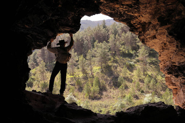 Mountain mannen avonturier met Australische hoed en rugzak in een grot met een dennenbos op de achtergrond - Foto, afbeelding