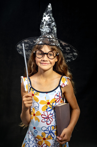 Girl sorceress, with a book and a magic wand, in a hat, on a black background - Photo, Image