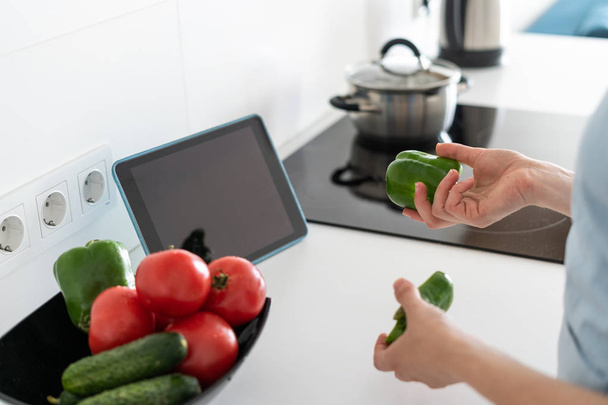 テーブルの上にデジタルタブレットで台所で食べ物を調理する女性, 手に野菜を保持 - 写真・画像