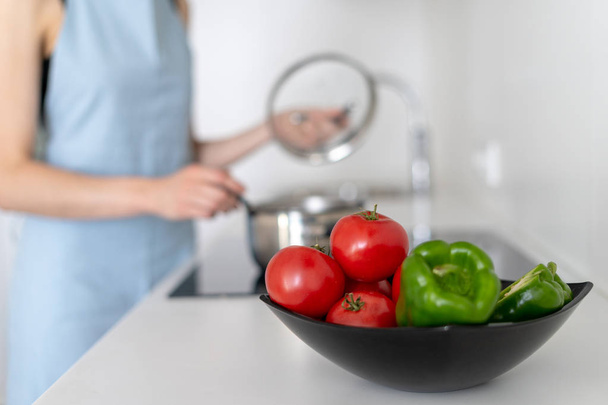 Tigela com legumes em fundo desfocado com mulher cozinhar alimentos, usando fogão elétrico
 - Foto, Imagem