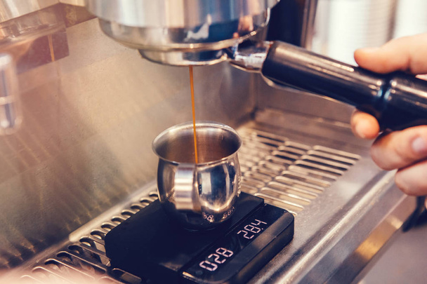 Close-up van professionele cappuccino espressomachine koffie maken. Dunne hete drank vloeibare stroom stroom komt gieten naar metalen mok kruik staan op schaal. Afgezwakt met warme gele film filters. - Foto, afbeelding