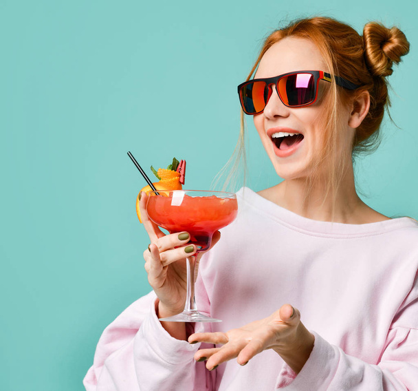 Joyful blonde girl in modern red sunglasses with tropic cocktail strawberry margarita has fun chatting with friends gesturing - Foto, Bild