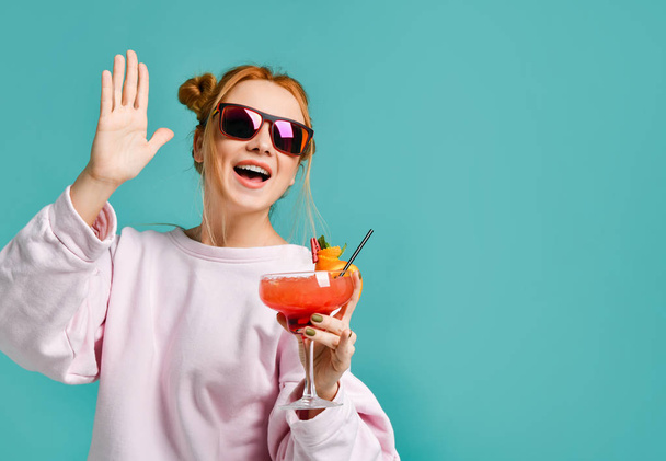Happy smiling clubbing young woman in modern red sunglasses with tropic cocktail in her hand greeting her friends - Foto, Bild