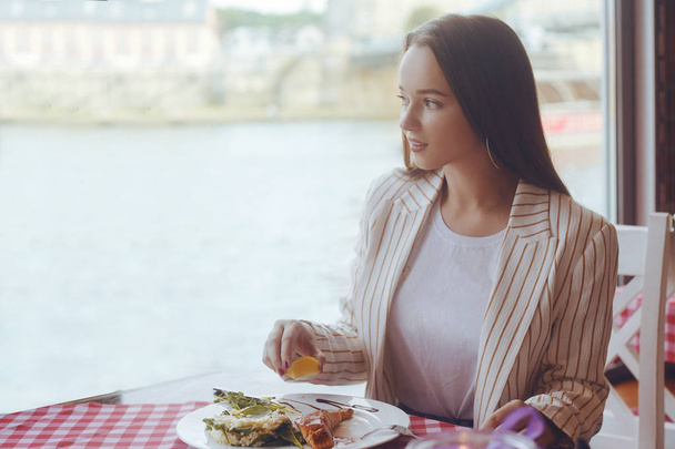 Brunette girl with long hair in a restaurant on ship on the background of the river. Girl tries salmon with rice. Girl eats appliances. The girl in the jacket after work eats.  - Photo, Image