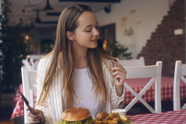 Brunette girl with long hair in a restaurant appetizing eating potatoes and burger. Girl tries burger and fries. Girl eats appliances. Woman smile in the jacket after work eats or stardy. Fast food. - Photo, Image
