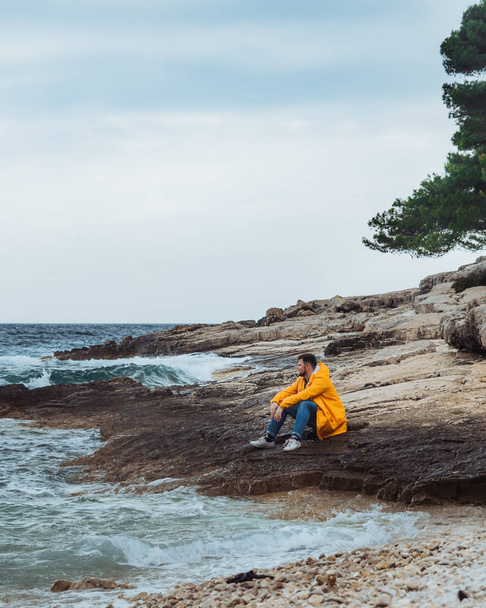 uomo seduto in impermeabile giallo sulla spiaggia rocciosa guardando il tempo tempestoso
 - Foto, immagini