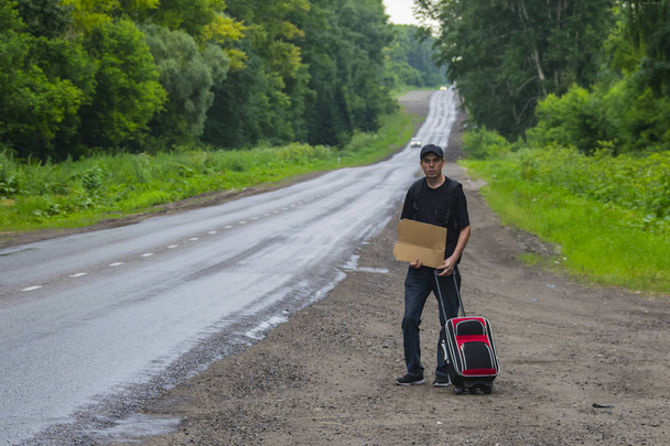 Un hombre con una maleta está cerca de la carretera. Él tiene un cartón en sus manos y quiere detener el coche
. - Foto, imagen