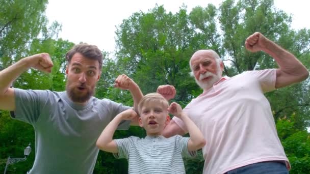 Portrait og happy family - grandpa, father and his son smiling and showing their muscles outdoor in park on background. Three different generation concept. - Footage, Video