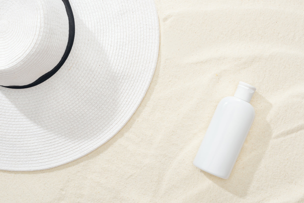 Top uitzicht van witte zonnebrandcrème en stro hoed op zand - Foto, afbeelding