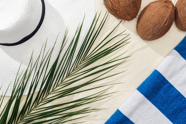 ylhäältä näkymä palmunlehtiä, raidallinen pyyhe, kookospähkinät ja valkoinen olki hattu hiekalla
 - Valokuva, kuva