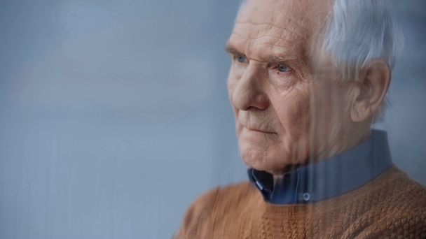 雨を降らせてカメラを見ながら窓の近くに立っている引退した男の選択的な焦点  - 映像、動画