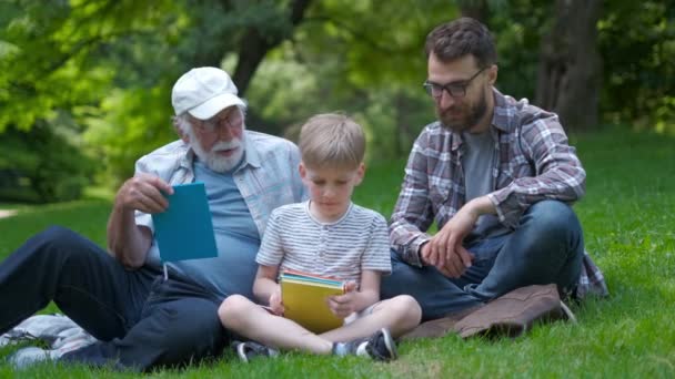 3世代の幸せな家族 - 本と公園の草の上に座っている父、祖父とブロンドの息子は、学校の準備をしながら読むことを学びます。彼らは笑い、楽しい、一緒に楽しい時間を過ごす - 映像、動画