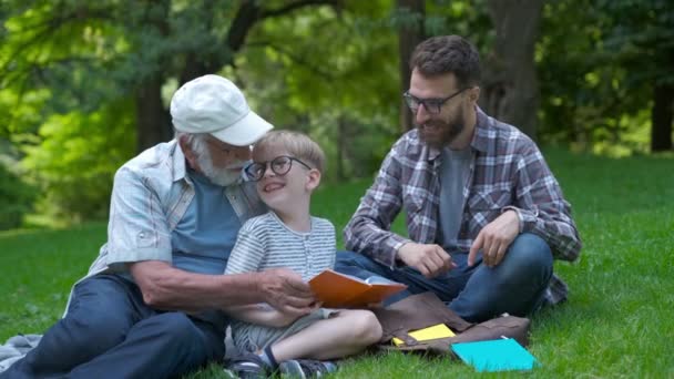 3世代の幸せな家族 - 本と公園の草の上に座っている父、祖父とブロンドの息子は、学校の準備をしながら読むことを学びます。彼らは笑い、楽しい、一緒に楽しい時間を過ごす - 映像、動画