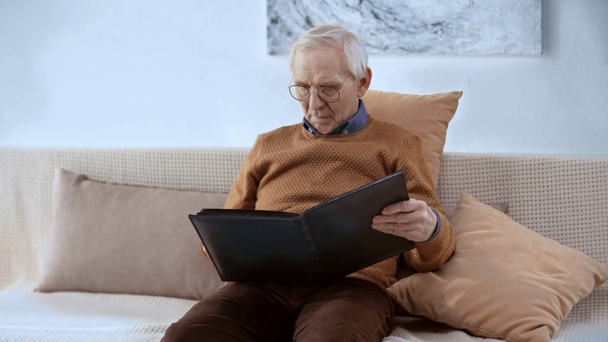 cheerful pensioner in glasses holding photo album, opening, looking at smiling at home  - Footage, Video