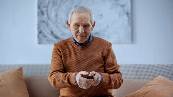 Senior Man ül a kanapén, és gazdaság távirányító nézése közben TV otthon  - Felvétel, videó
