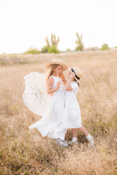 Cute little girls sisters with blond  hair in a summer field at sunset in white dresses with a straw hat - Фото, изображение