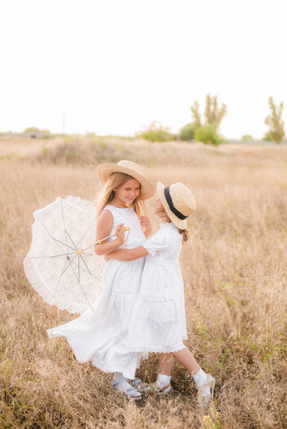 Cute little girls sisters with blond  hair in a summer field at sunset in white dresses with a straw hat - Φωτογραφία, εικόνα