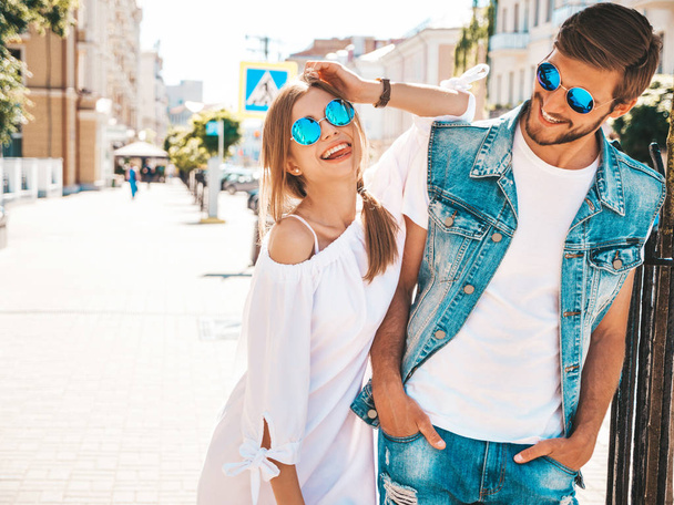Smiling beautiful girl and her handsome boyfriend posing in the street. Woman in casual summer dress and man in jeans clothes. Happy cheerful couple family having fun in sunglasses - Photo, image