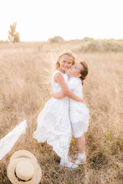 Cute little girls sisters with blond  hair in a summer field at sunset in white dresses with a straw hat - Zdjęcie, obraz