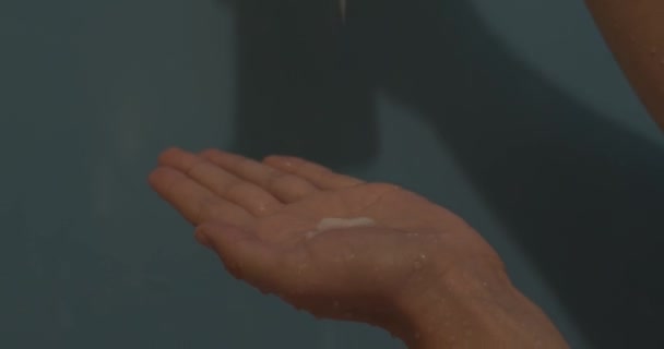 Mujer verter champú, bálsamo, máscara, crema, líquido cosmético en la mano en la ducha. Mano con gotas de agua usando líquido cosmético sobre fondo azul de cerca
 - Metraje, vídeo