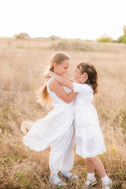 Cute little girls sisters with blond  hair in a summer field at sunset in white dresses with a straw hat - Fotó, kép