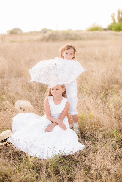 Cute little girls sisters with blond  hair in a summer field at sunset in white dresses with a straw hat - Photo, image