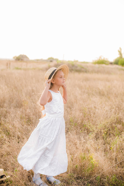 Cute little girl with blond hair in a summer field at sunset with a white dress with a straw hat - Photo, image
