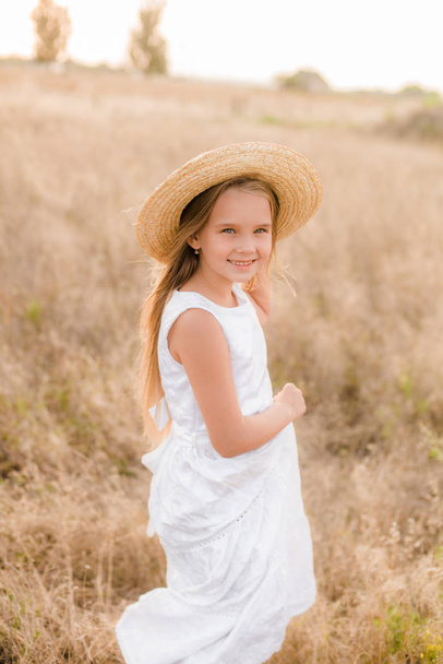Cute little girl with blond hair in a summer field at sunset with a white dress with a straw hat - Fotó, kép