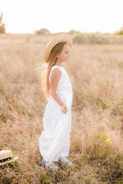 Cute little girl with blond hair in a summer field at sunset with a white dress with a straw hat - Фото, изображение