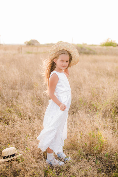 Cute little girl with blond hair in a summer field at sunset with a white dress with a straw hat - Foto, Imagem