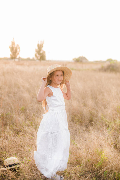 Cute little girl with blond hair in a summer field at sunset with a white dress with a straw hat - Foto, Imagen