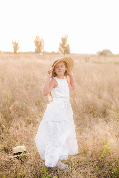 Cute little girl with blond hair in a summer field at sunset with a white dress with a straw hat - Fotó, kép