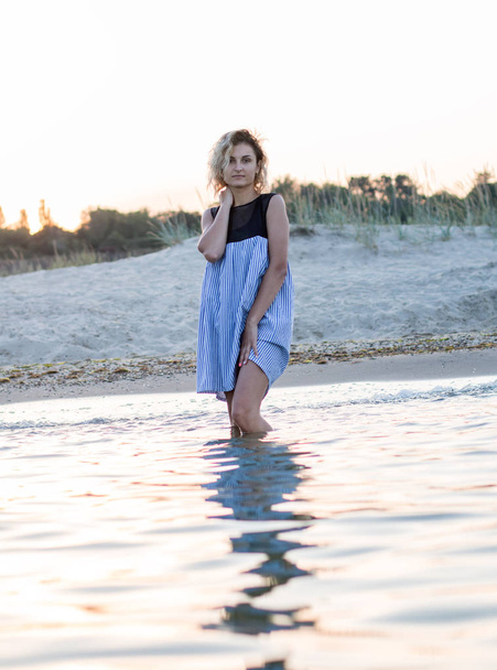 femmina con i capelli ricci in abito è in piedi in acqua sulla spiaggia, tramonto sullo sfondo
 - Foto, immagini