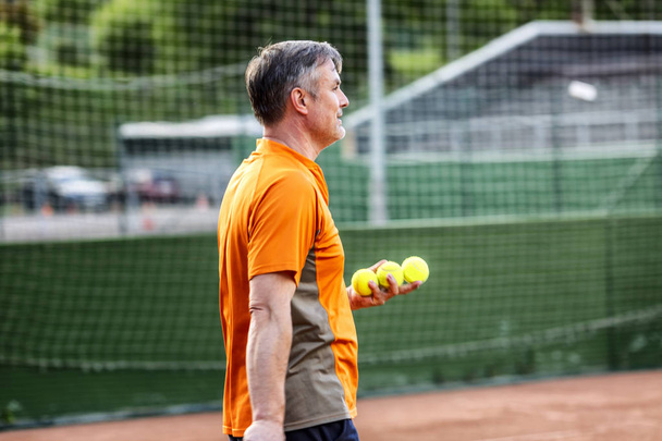 A middle-aged man plays tennis on a court with a natural earth surface on a sunny summer day.  - Zdjęcie, obraz